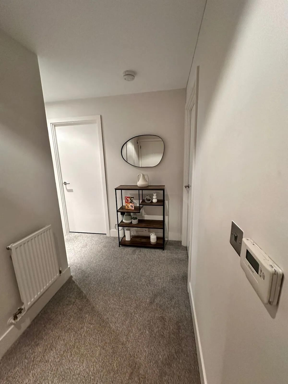 A hallway with a white door, a radiator, a small shelf with items, and a wall-mounted mirror.