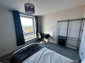 A bedroom with a bed, a desk, a chair, a black metal shelf unit, a window with curtains, and a hanging light fixture. - Cosy, Comfy 2 Bed Apartment Canary Wharf (London)