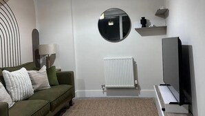 Un salon moderne avec un canapé vert, un miroir rond, un téléviseur à écran plat, un radiateur et un luminaire suspendu.