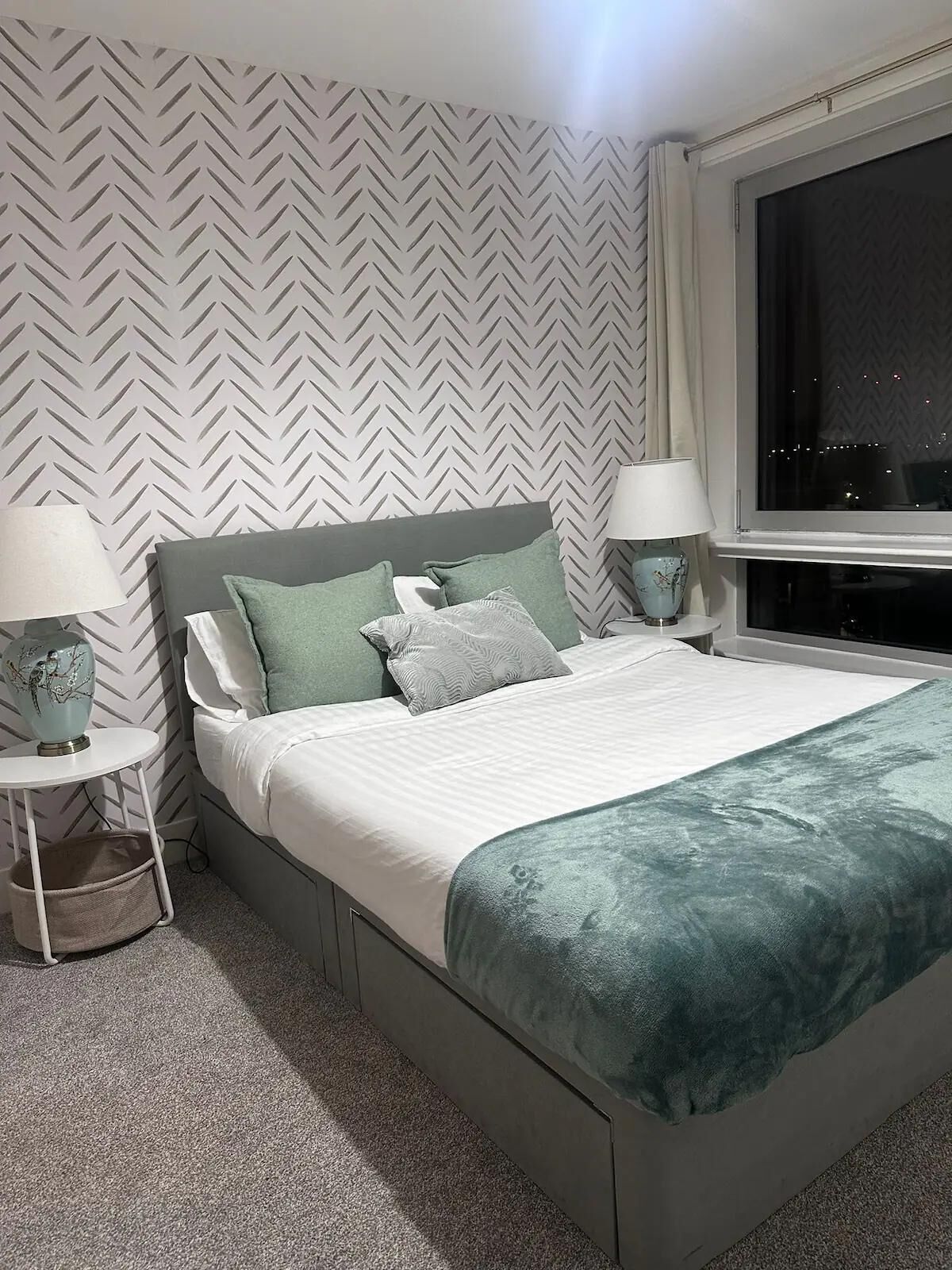 Une chambre moderne avec un lit gris, du linge de lit blanc et un plaid turquoise. On y trouve deux tables de chevet avec des lampes, une fenêtre donnant sur la ville la nuit, et un mur orné d’un motif en chevrons.
