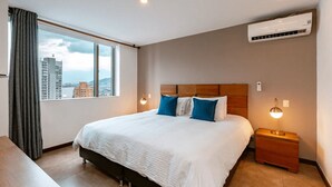 Una habitación de hotel moderna con una cama grande, cabecera de madera, mesita de noche con lámpara, unidad de aire acondicionado y vista a la ciudad a través del ventanal.