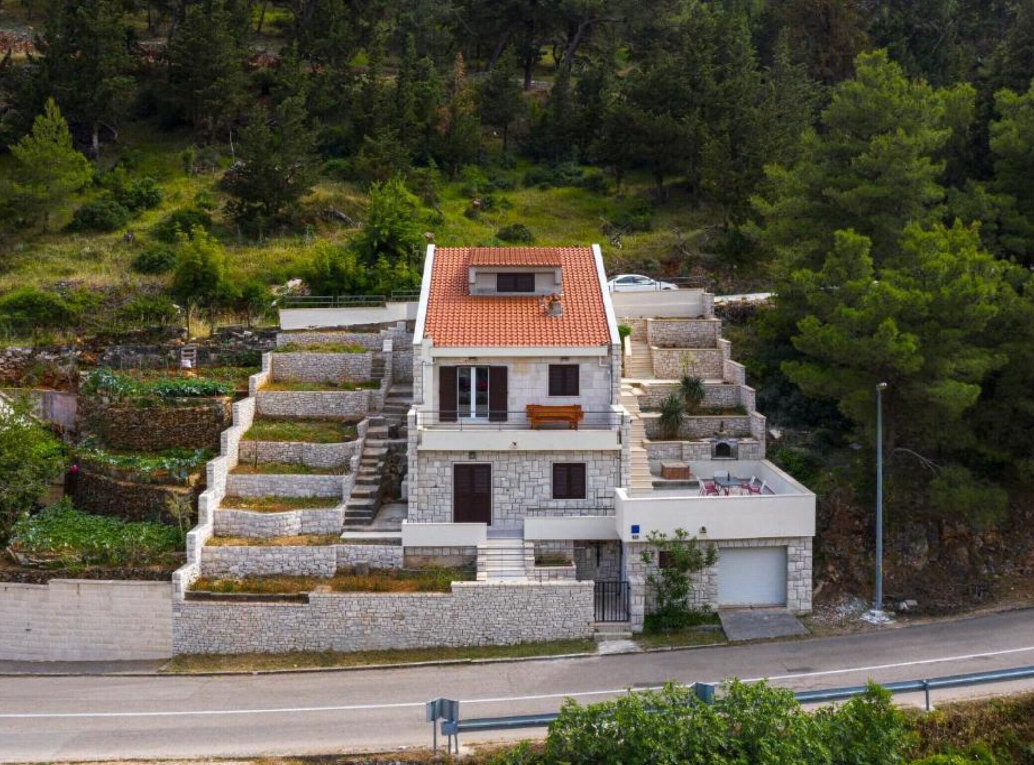 A modern two-story house with a stone facade and a red-tiled roof, surrounded by lush greenery and a hillside.