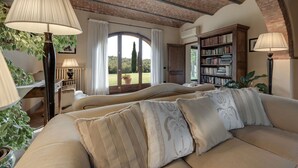 Ein gemütliches Wohnzimmer mit Ziegeldecke, einem großen Fenster mit Vorhängen, einer Couch mit gemusterten Kissen, einem Bücherregal und einer Lampe.