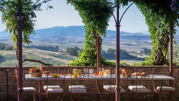 An outdoor dining setup with a table and chairs, surrounded by greenery and a scenic view of hills.