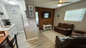 Una sala de estar moderna con un sofá de cuero marrón, una mesa de comedor de madera con sillas, una zona de cocina con armarios blancos, un televisor de pantalla plana montado en la pared y un ventilador de techo.
