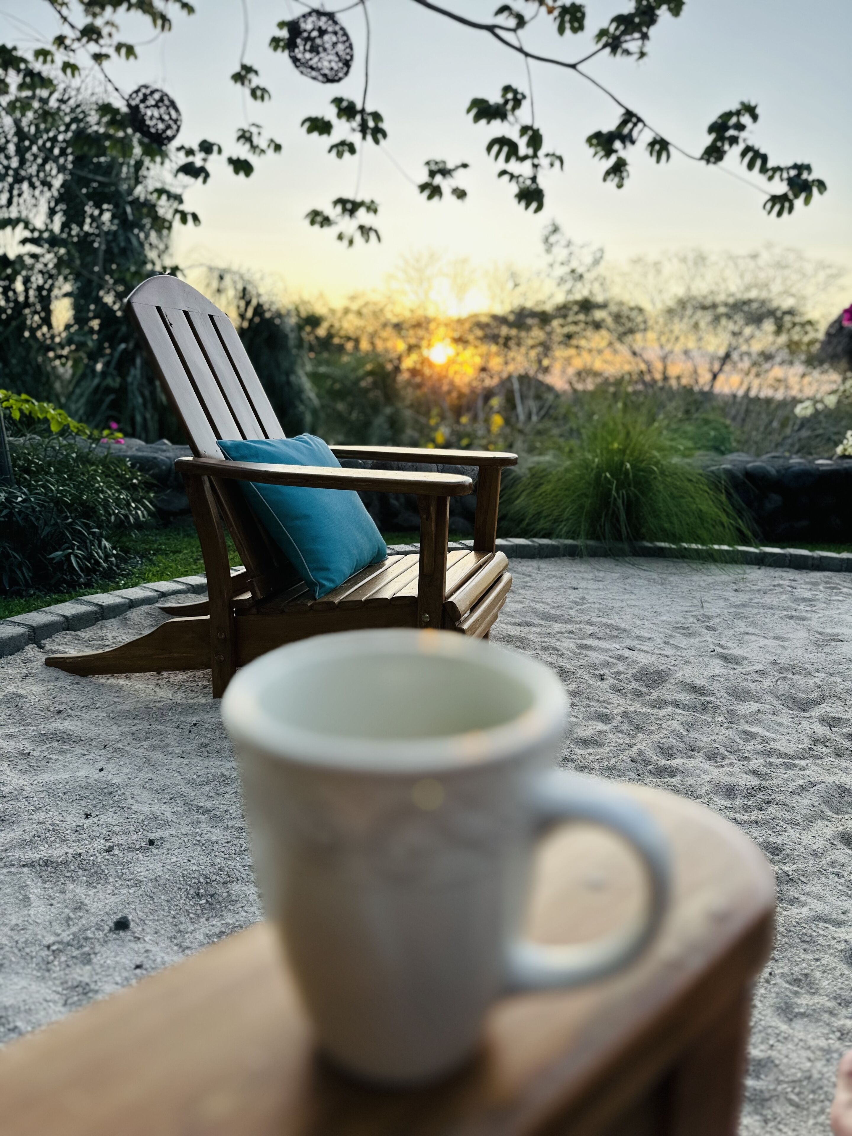 A wooden chair with a cushion and a mug on a stand, set on a gravel surface with a sunset in the background.