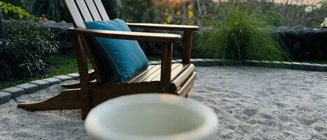A wooden chair with a cushion and a mug on a stand, set on a gravel surface with a sunset in the background.