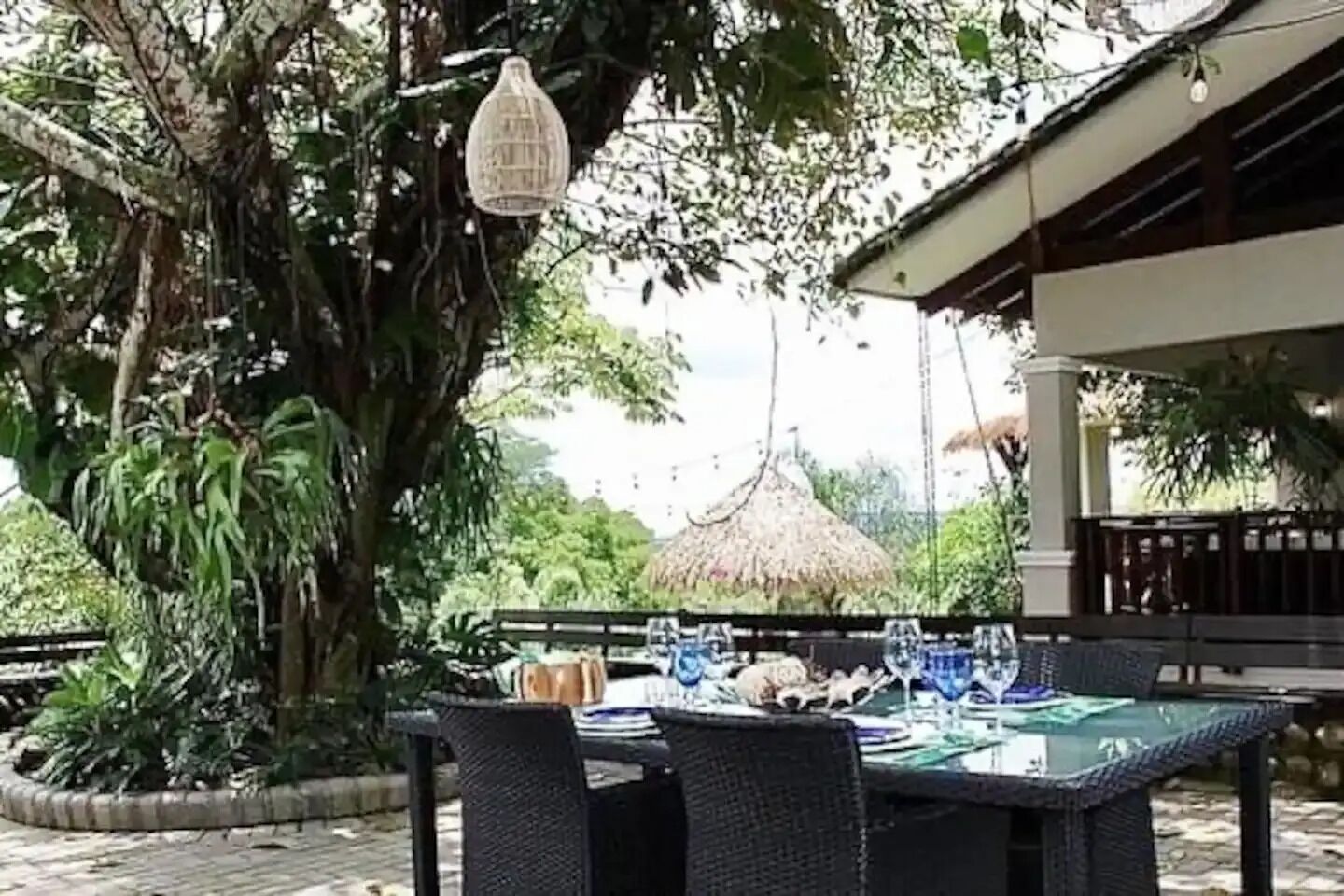 An outdoor dining area with a table set for two, surrounded by lush greenery and a thatched-roof structure in the background.