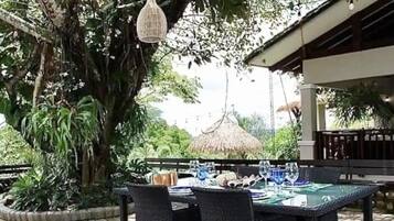 An outdoor dining area with a table set for two, surrounded by lush greenery and a thatched-roof structure in the background.