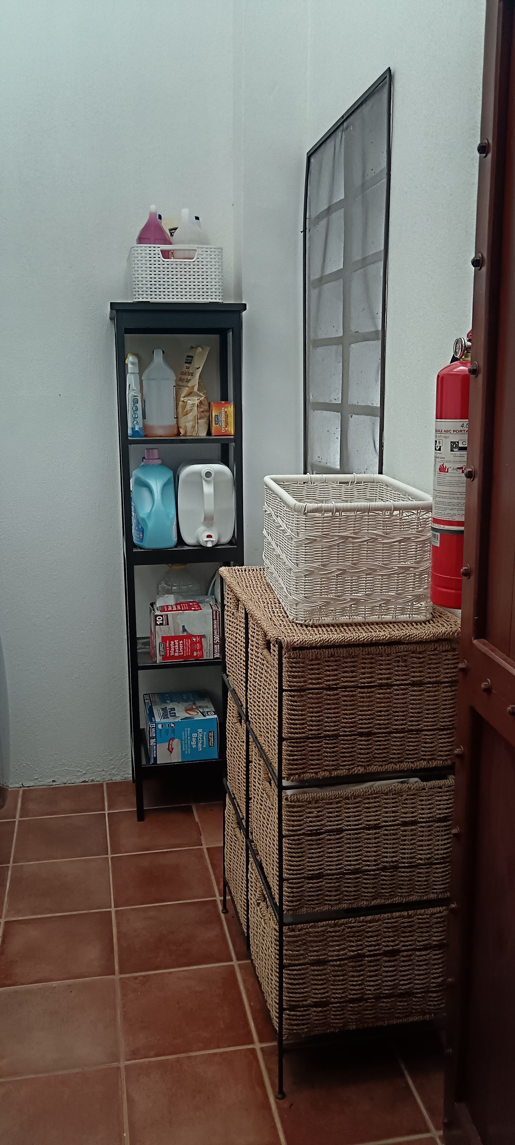 Rincón de una habitación con un estante que contiene productos de limpieza, una cesta de mimbre y un extintor.