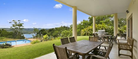 Un patio exterior con mesa y sillas de madera, una parrilla y vistas a una piscina y follaje.