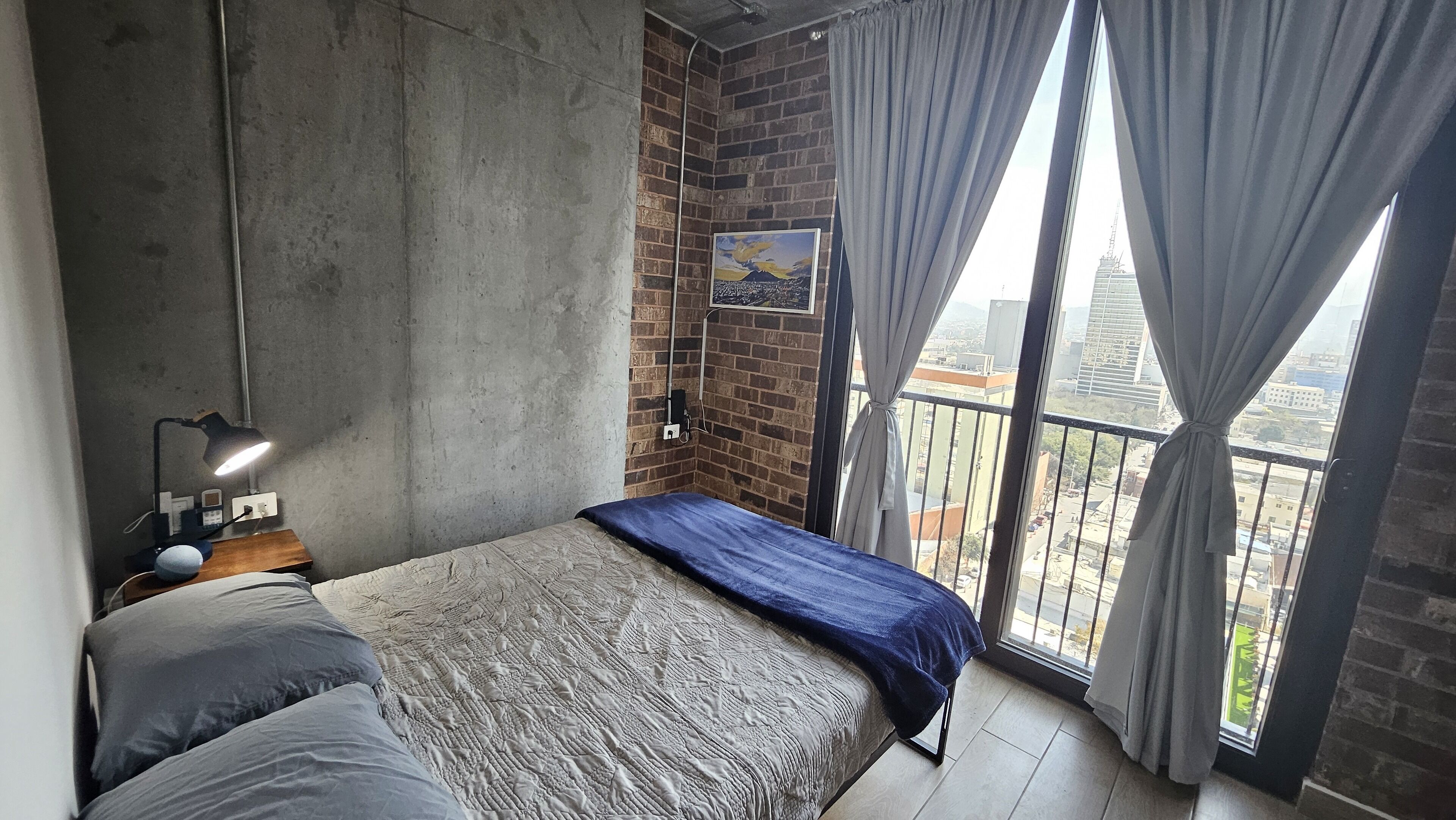 A bedroom with a concrete wall, a bed with a blue blanket, a desk with a lamp, and a view of the city through large windows.