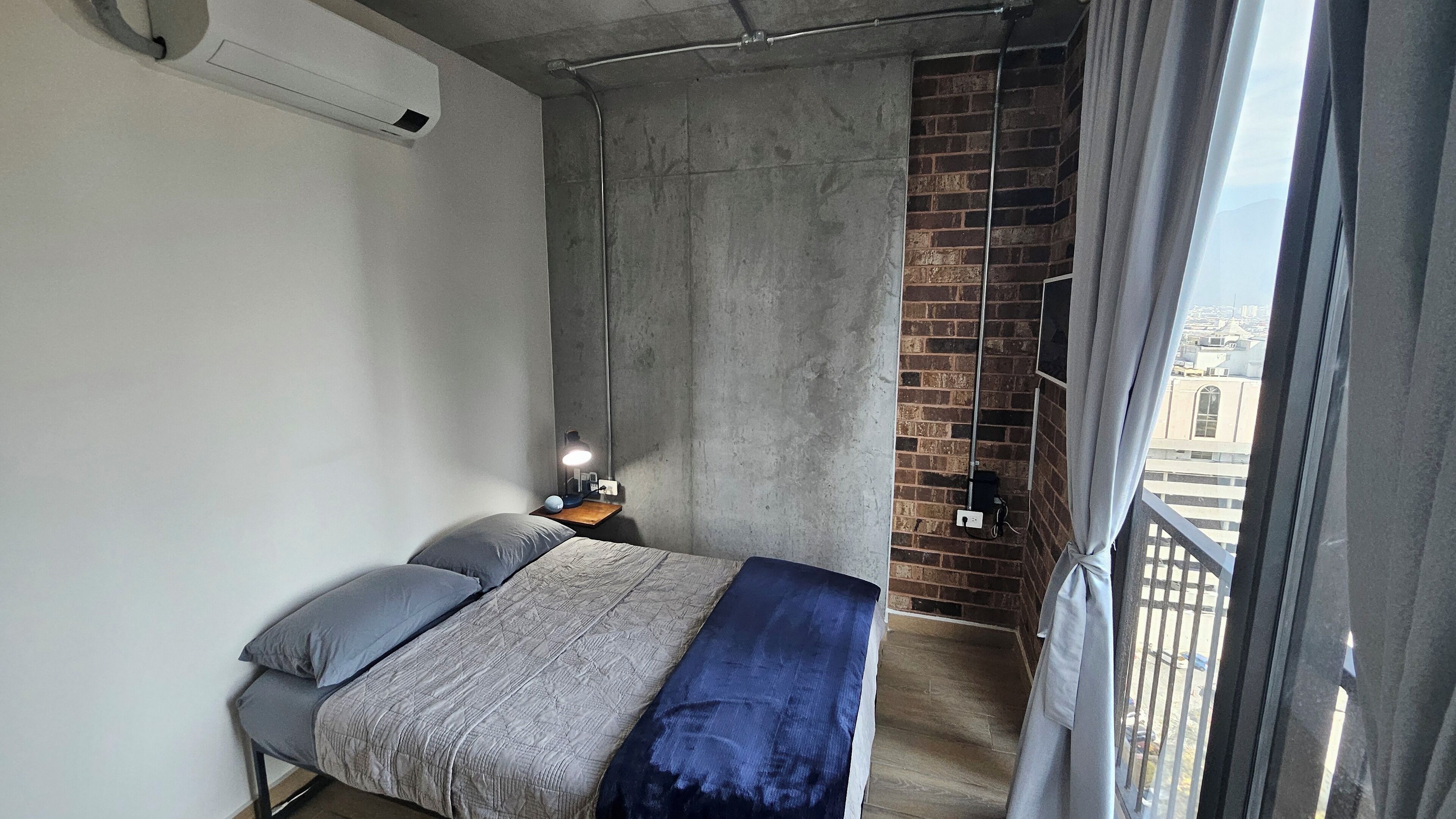 A bedroom with a bed, a nightstand, a lamp, a window with a view of buildings, and a brick wall.