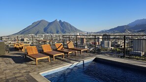 Un área de piscina en la azotea con sillones de madera y vistas a un paisaje urbano y montañas.