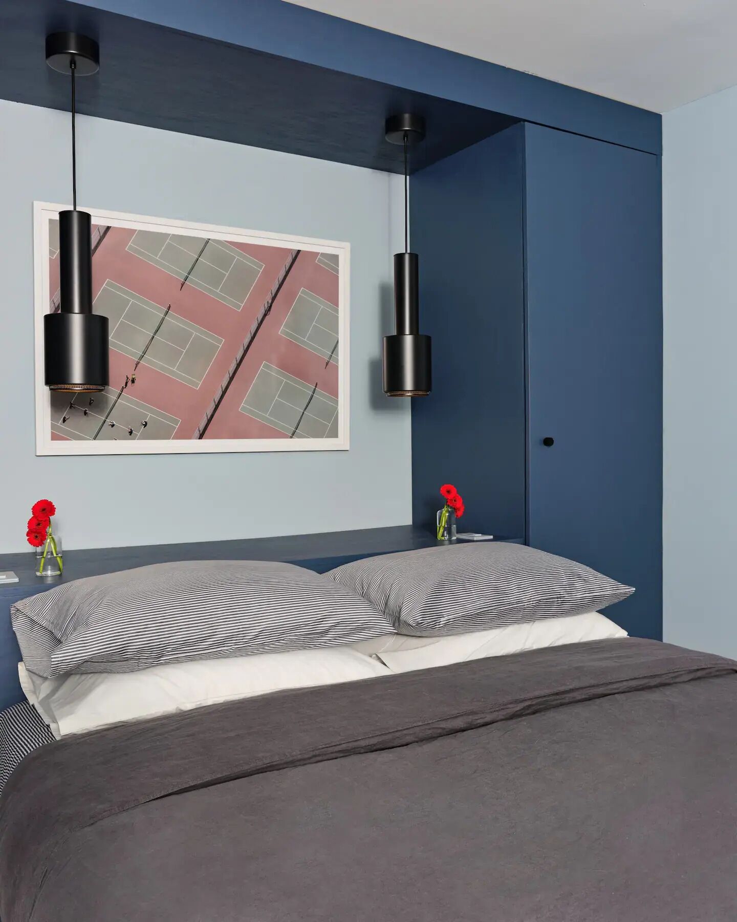 Een moderne slaapkamer met een bed, twee zwarte hanglampen, een foto van een tennisbaan in een lijst en twee rode bloemen op een nachtkastje.