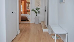 A hallway with a wooden floor, white walls, a white bench, and a door leading to a bedroom.
