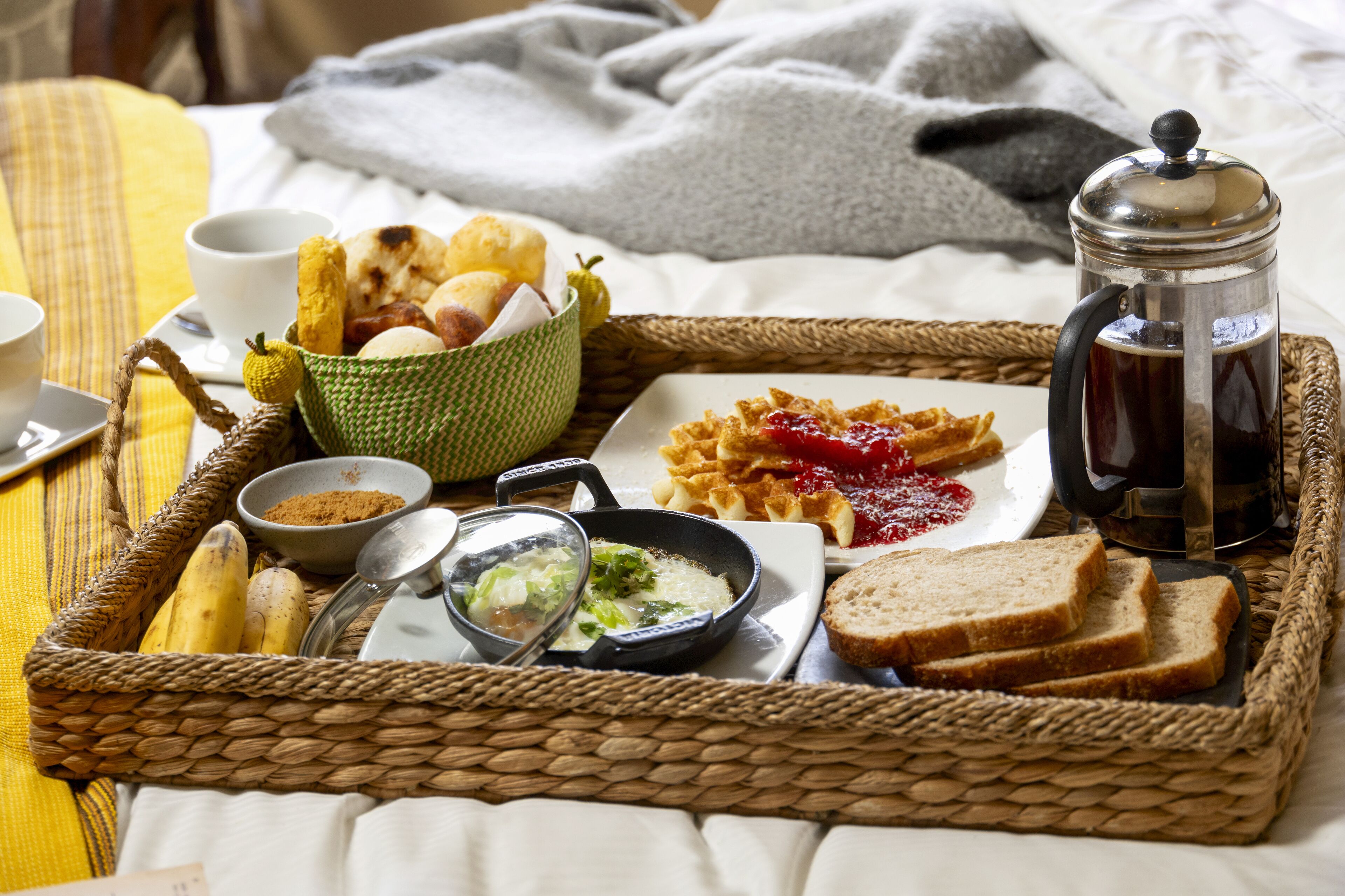 A breakfast tray with bread, jam, soup, and a coffee pot on a bed.
