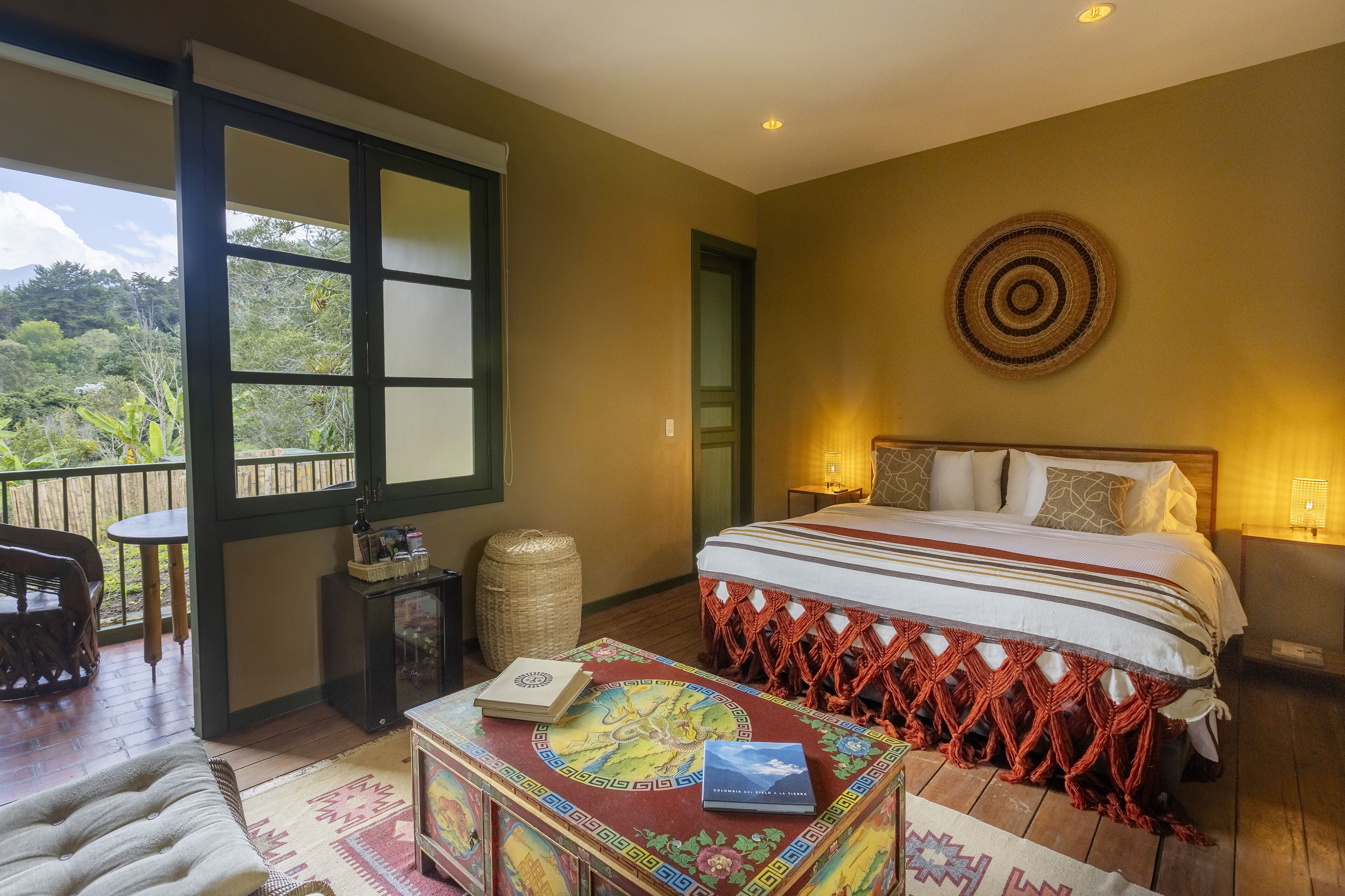 A bedroom with a large bed, a small table with books, a decorative box, and a view of the outdoors.