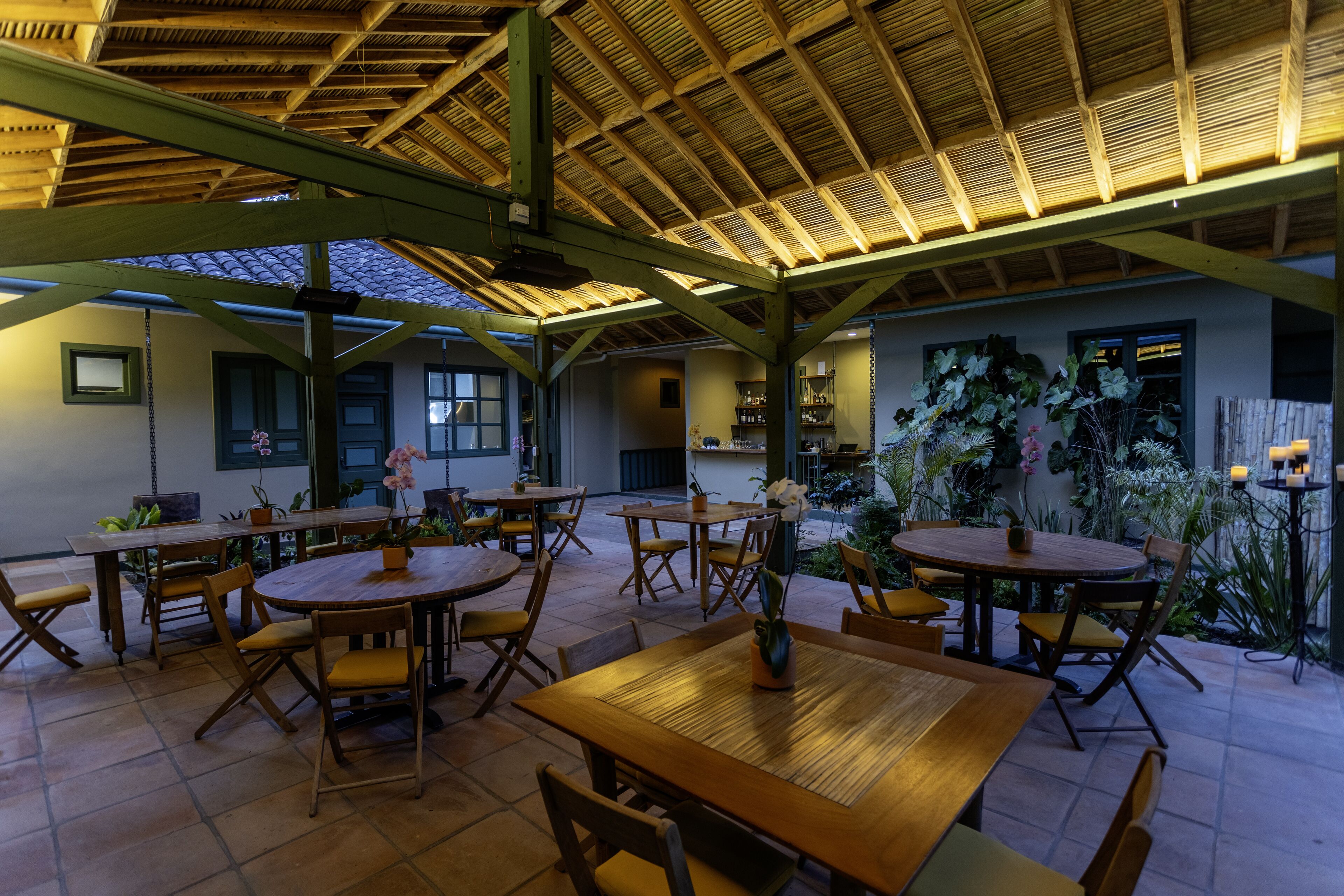 An outdoor dining area with wooden tables and chairs, surrounded by potted plants and a thatched roof structure.