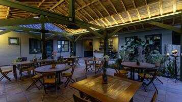 An outdoor dining area with wooden tables and chairs, surrounded by potted plants and a thatched roof structure.