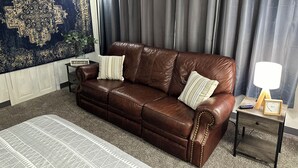 Un canapé en cuir marron avec deux coussins, une petite table d’appoint en bois avec une lampe, et un grand tapis à motifs.