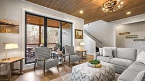 A spacious living room with a large gray sofa, a wooden coffee table, and a fireplace. There is a staircase leading to an upper level, and a large window providing a view of the outdoors.