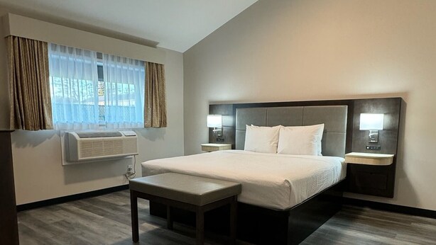 Una habitación de hotel moderna con una cama grande, mesitas de noche, un banco y un ventanal con cortinas.