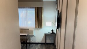 Un pasillo con piso de madera que lleva a una habitación con cama, una ventana con cortinas y un escritorio con lámpara.