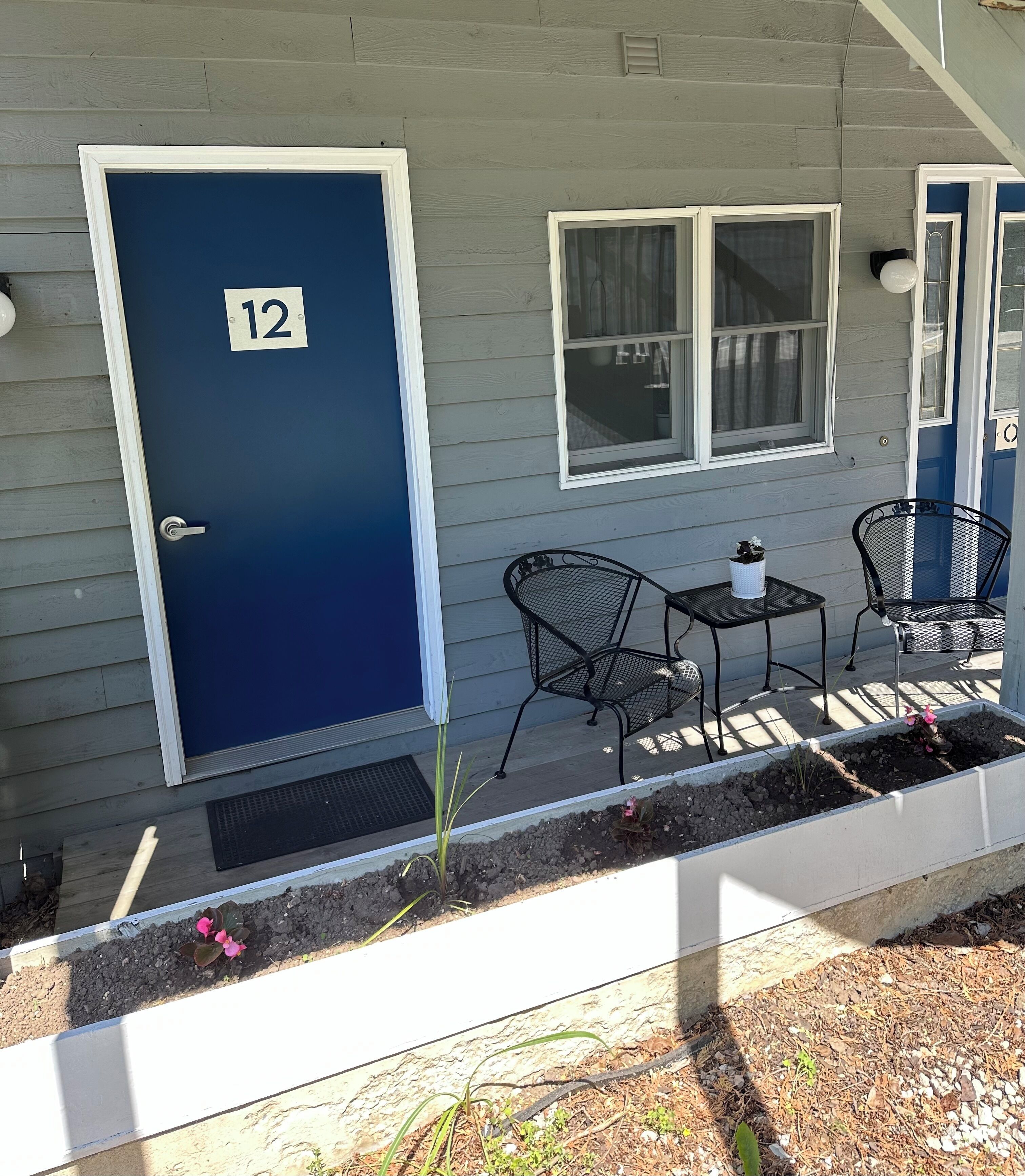 Une porte bleue portant le numéro 12, une petite table, deux chaises et une fenêtre avec des rideaux.