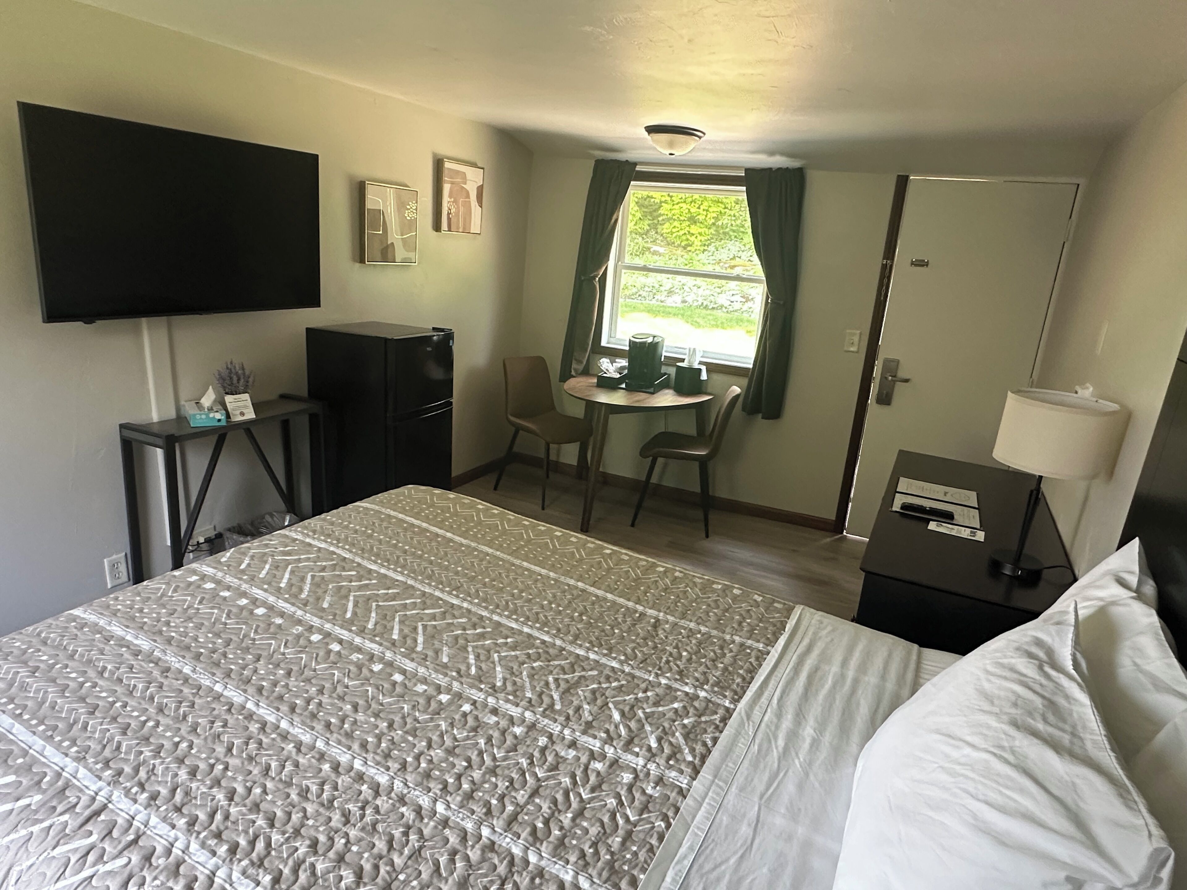 Una habitación de hotel con cama, escritorio con lámpara, televisor, refrigerador, ventana con cortinas y puerta.