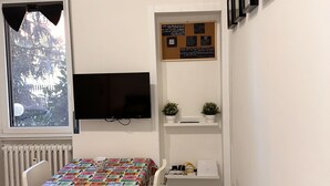 Una habitación pequeña con una mesa cubierta por un mantel colorido, un televisor montado en la pared, un tablón de anuncios y una ventana que da a un paisaje de árboles.