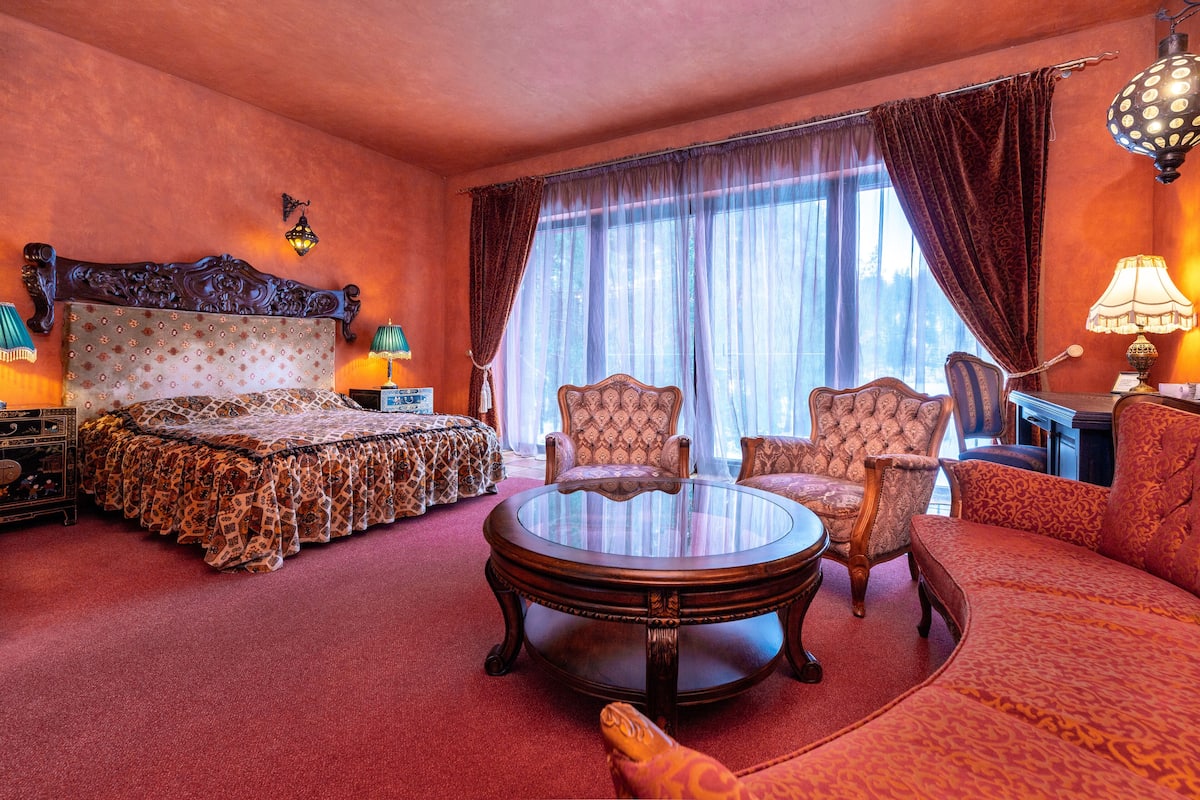 A room with a large bed, two armchairs, a round table, and a desk with a lamp.