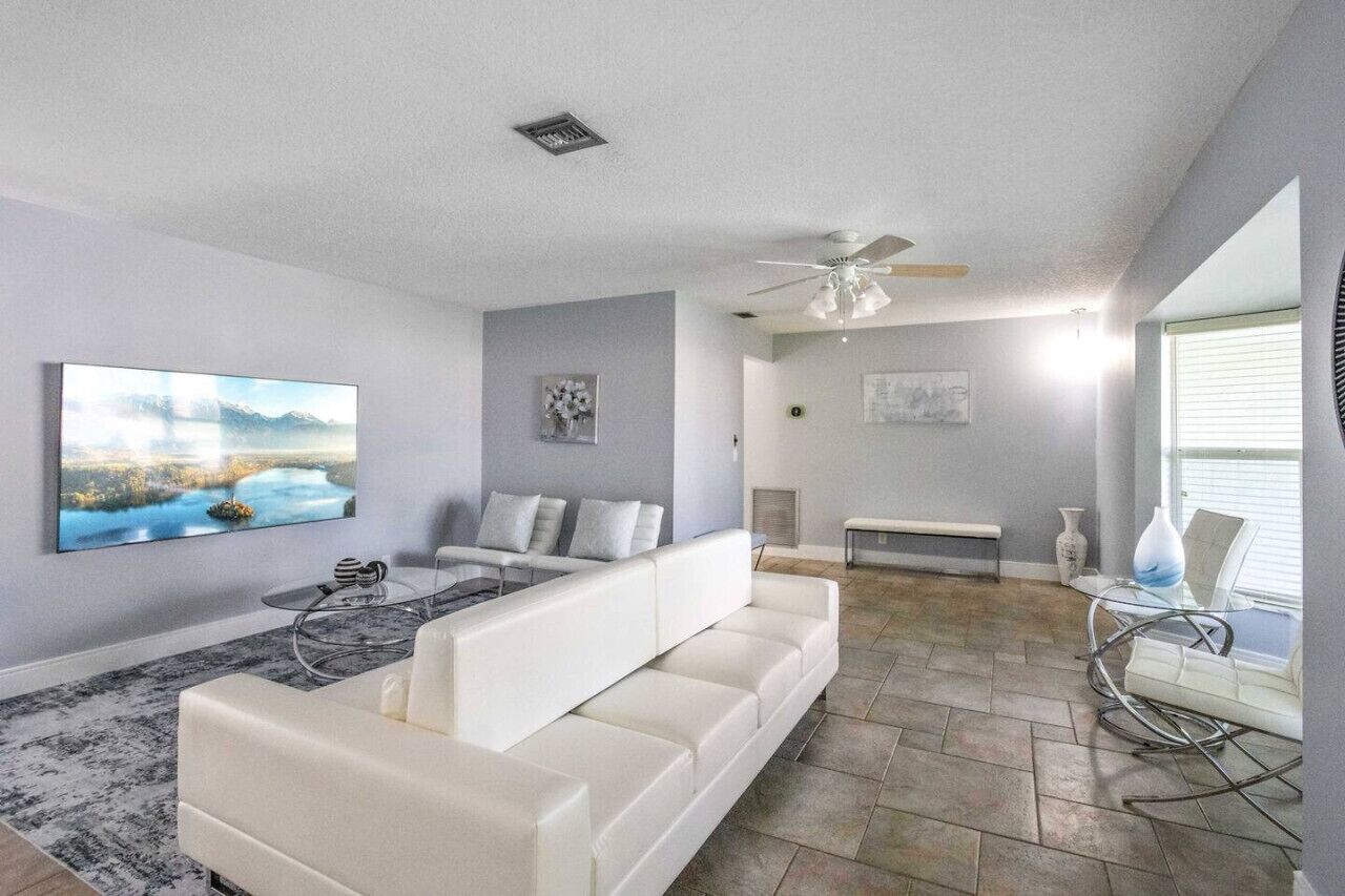 A modern living room with a white sofa, a coffee table, and a ceiling fan. There is a painting on the wall and a window with blinds.