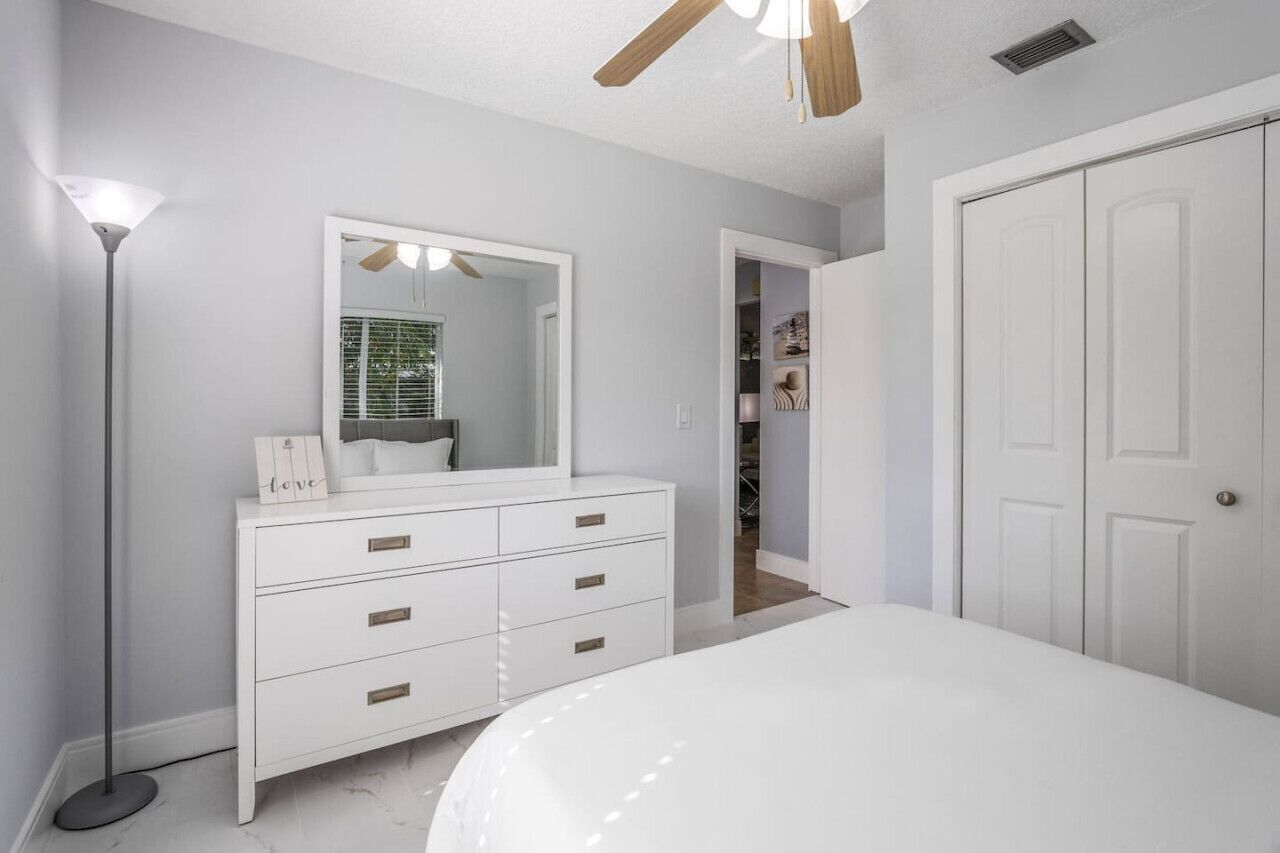 Une chambre avec une commode blanche, un grand miroir, un ventilateur de plafond et un placard avec une porte.