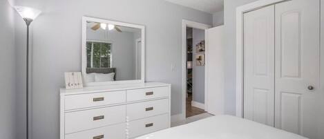 Une chambre avec une commode blanche, un grand miroir, un ventilateur de plafond et un placard avec une porte.