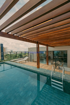 Piscina en la azotea con una pérgola de madera, sillas de descanso y barandilla de vidrio con vistas a la ciudad.