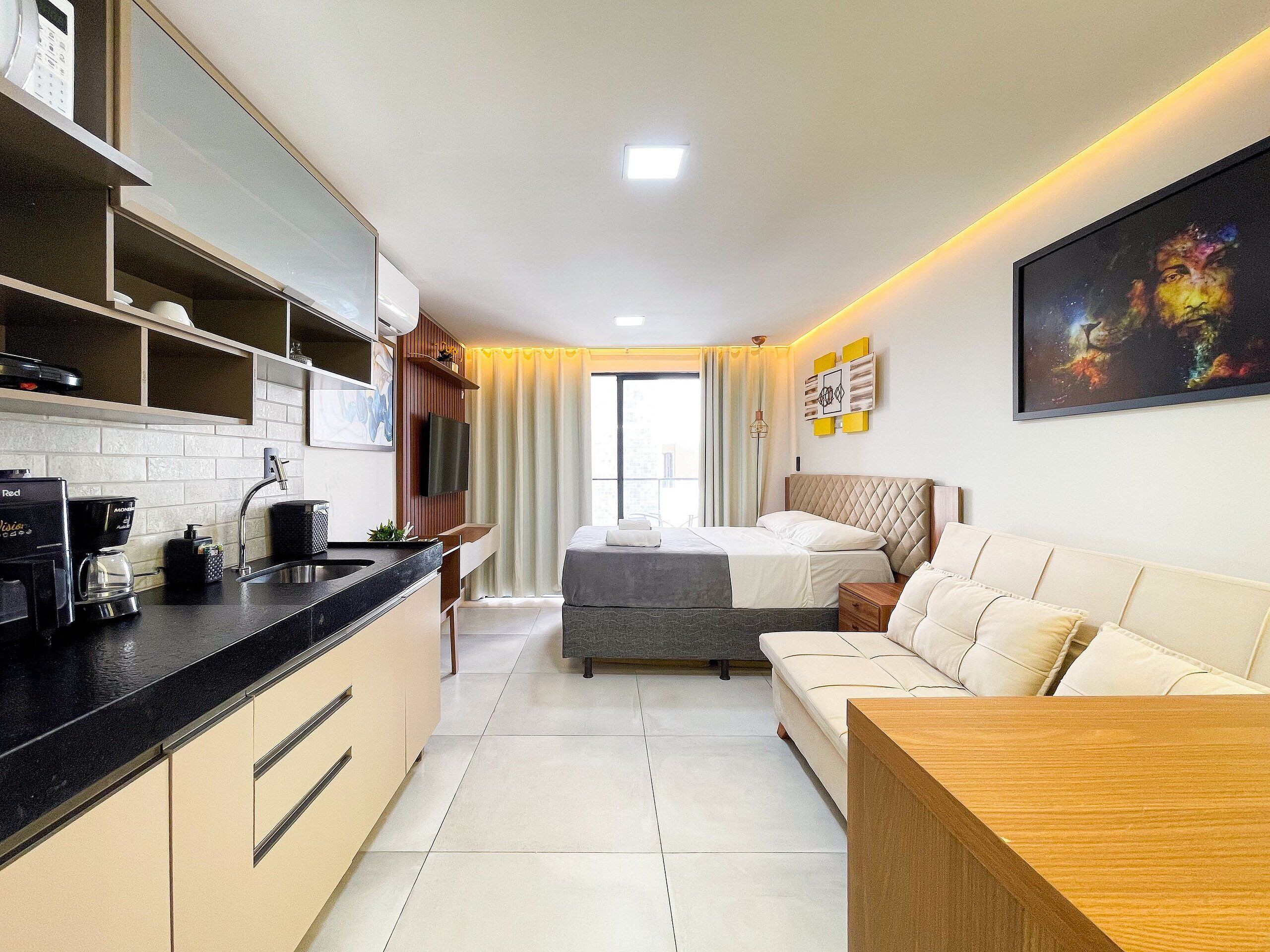 A modern hotel room with a kitchenette, a bed with a grey headboard, a sofa, and a large painting on the wall.