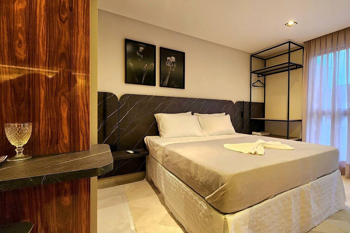 A modern hotel room with a large bed, a bedside table, a nightstand, a wardrobe, and a large window with curtains.