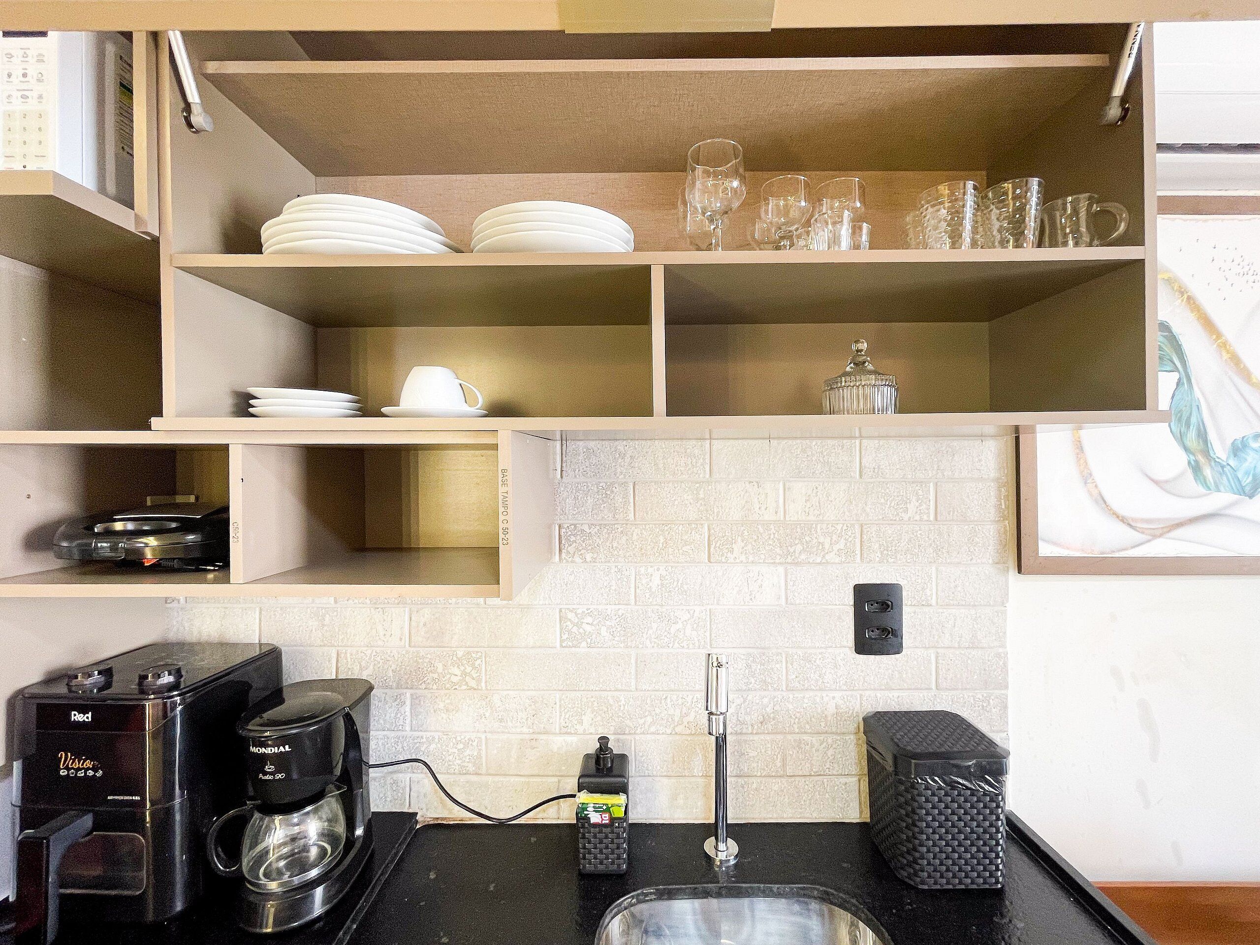 Una cocina con cafetera, pileta y estantes con platos y vasos.