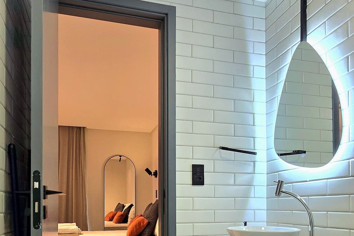 A modern bathroom with a white sink, a mirror, and a bed visible through an open door.