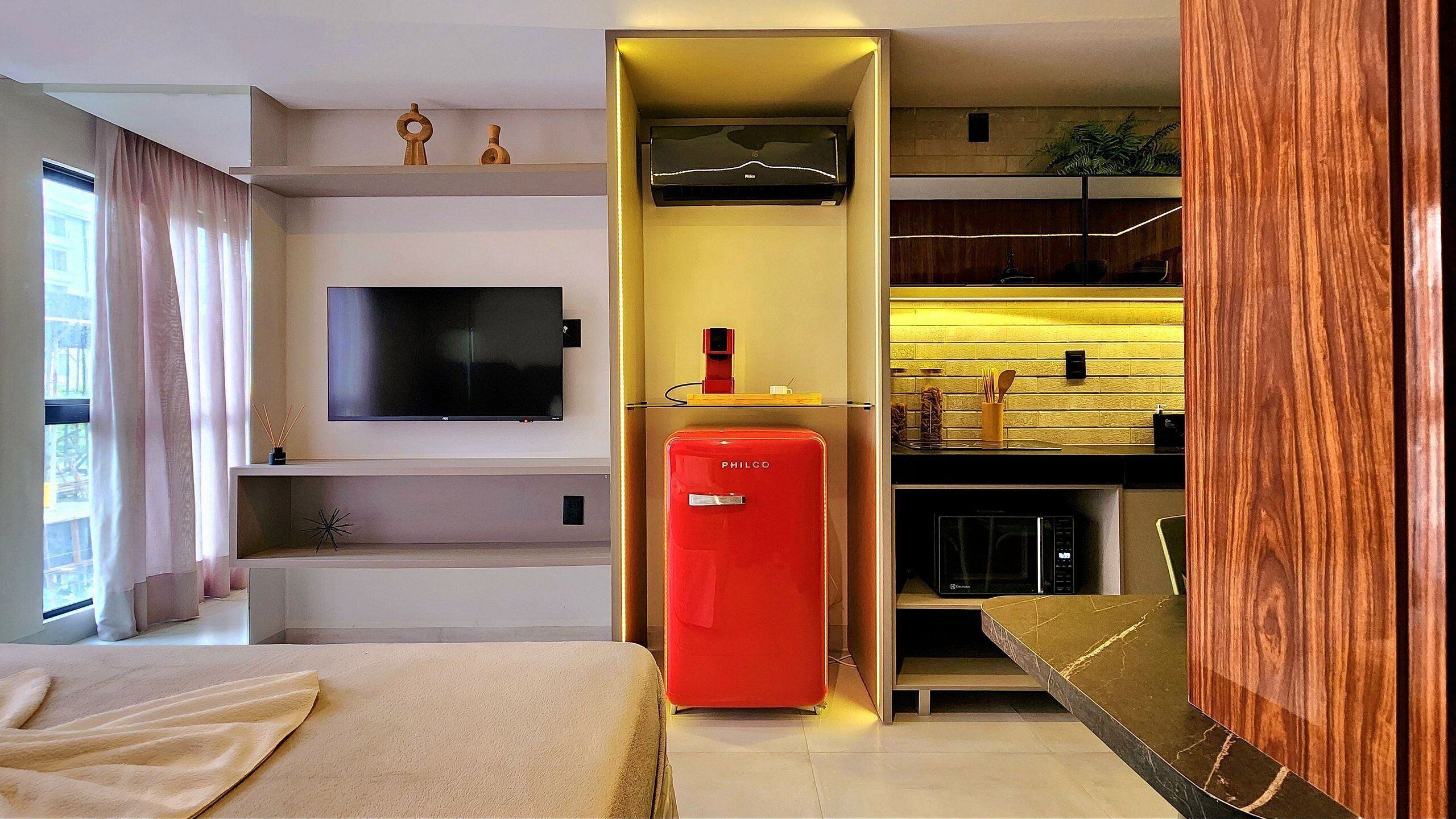 Ein modernes Hotelzimmer mit Flachbildfernseher, einer roten Minibar, einem Holzschrank und einer Küchenzeile mit Mikrowelle.