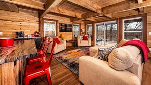 A rustic cabin interior with wooden walls, a dining area with red chairs, a sofa with a zebra pillow, and large windows. - 2BR, 1BA  HAND BUILT MOUNTAIN TOP TREE HOUSE with Hot tub (Nashville)