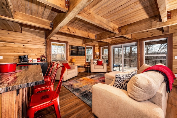 A rustic cabin interior with wooden walls, a dining area with red chairs, a sofa with a zebra pillow, and large windows.