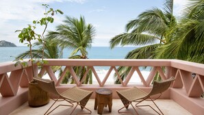 A balcony with wicker chairs and a small table, overlooking a beach with palm trees and the sea.