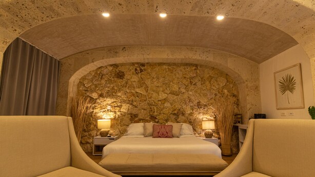 A bedroom with a stone wall, a bed with white linens, two bedside tables with lamps, and two armchairs.