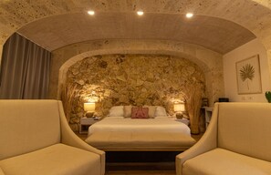 Une chambre avec un mur en pierre, un lit recouvert de draps blancs, deux tables de chevet avec des lampes et deux fauteuils.
