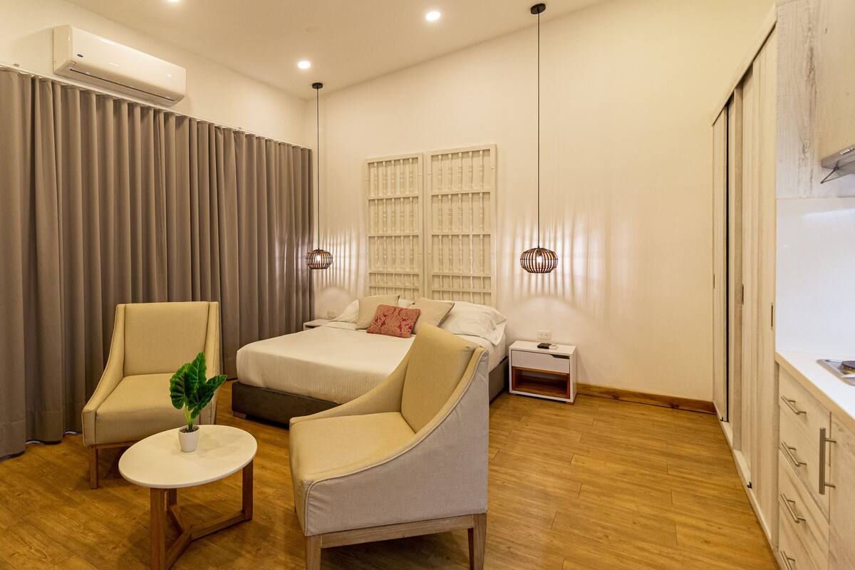 Una habitación de hotel moderna con cama, dos sillones, una mesita, aire acondicionado, una mesita de noche y una ventana con persianas.