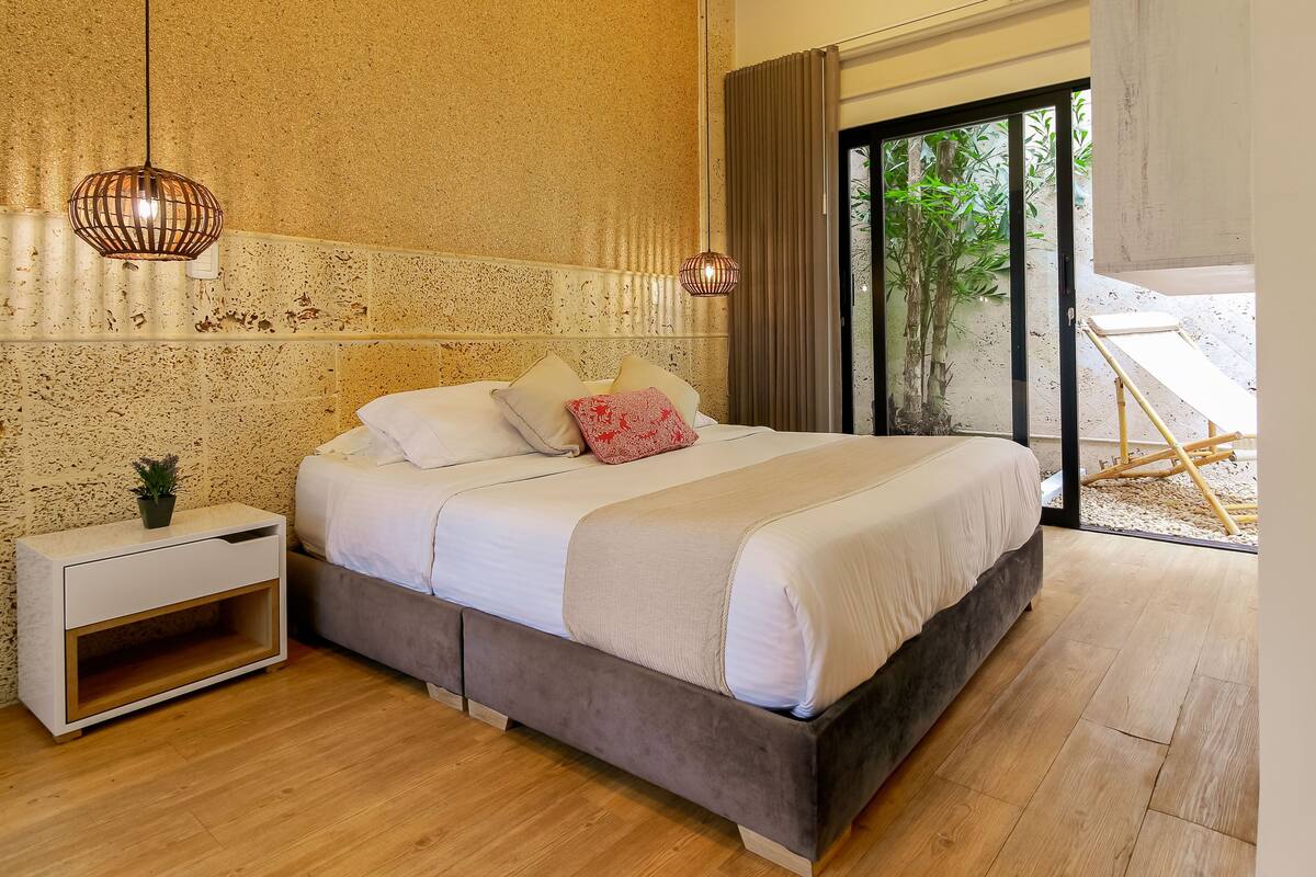 Un dormitorio con una cama grande, una mesita de noche, una planta en maceta y vistas al exterior con vegetación.