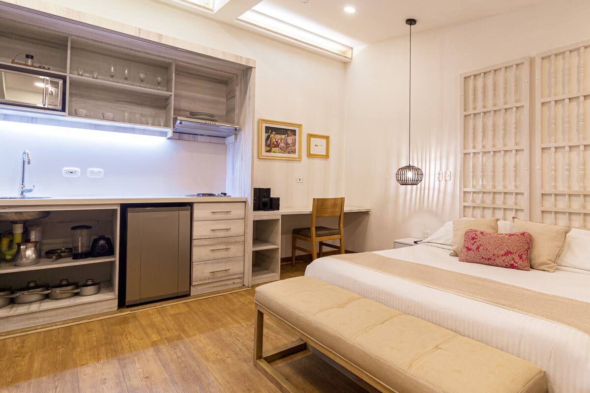Habitación de hotel compacta con kitchenette, una cama con un cojín decorativo, un banco y un escritorio con silla.