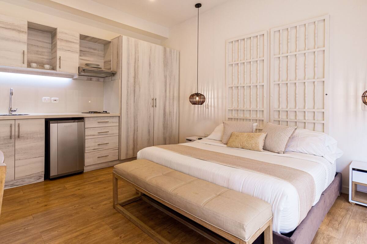 Un dormitorio moderno con una cama grande, mesitas de noche, un banco, un escritorio empotrado con microondas y un armario.